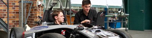 Two men looking at a laptop screen. One kneeling, the other sitting inside a race car.