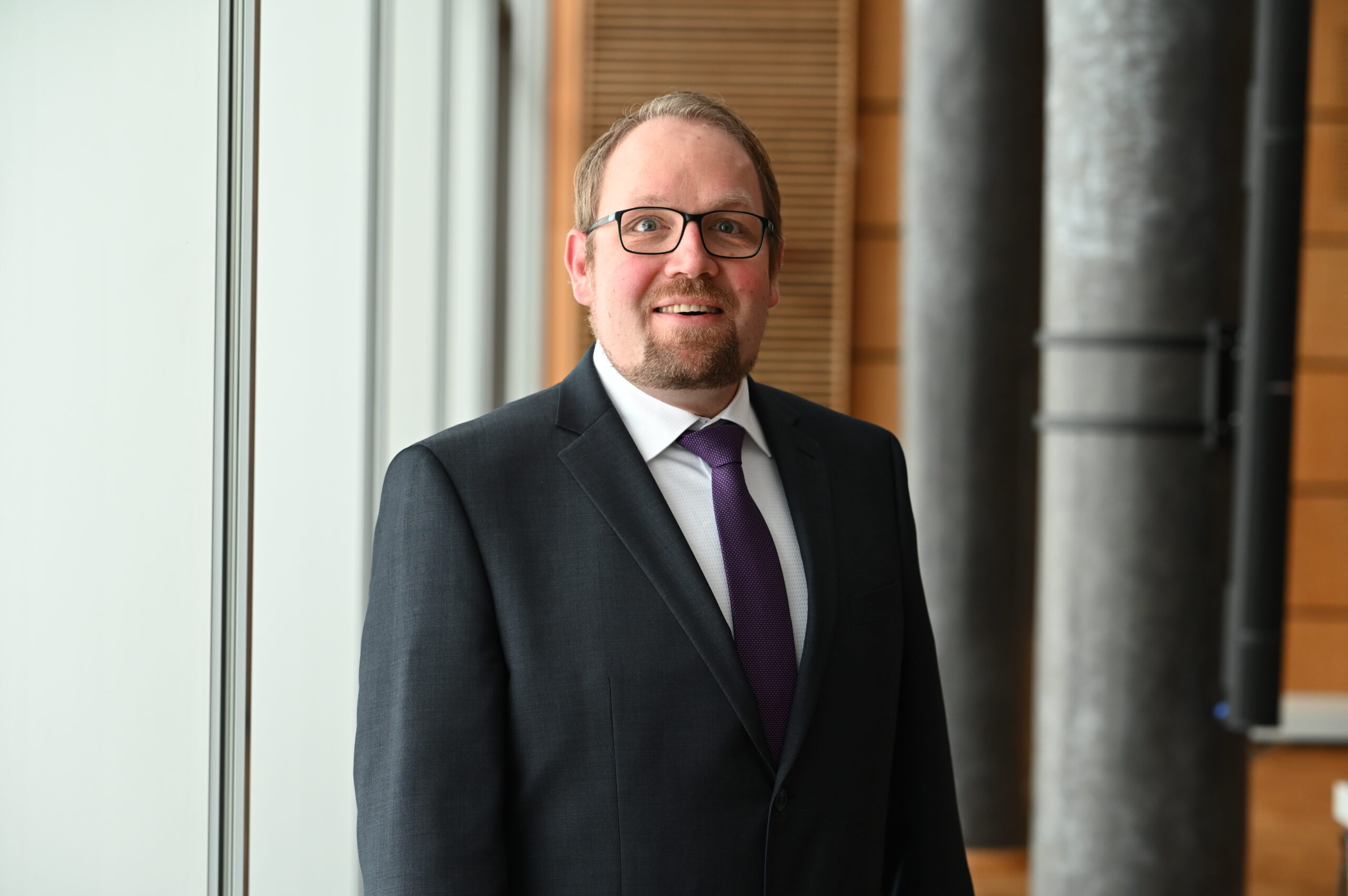 Ein Mann mit Brille, in einem schwarzen Anzug, einem weißen Hemd und einer violetten Krawatte steht drinnen vor großen Fenstern und grauen Säulen und lächelt in die Kamera – dabei strahlt er die selbstbewusste Haltung eines Kanzlers aus, der seine erste Bilanz vorstellt. at Hochschule Coburg