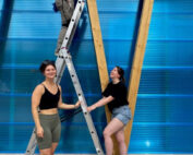 Drei Personen der Hochschule Coburg arbeiten gemeinsam an einem Bauprojekt. Einer steht auf einer Leiter, die anderen daneben. Sie bauen eine Holzkonstruktion mit einer blau durchscheinenden Wand zusammen. Die Szene spielt im Freien und sie wirken fröhlich.