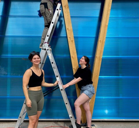 Drei Personen der Hochschule Coburg arbeiten gemeinsam an einem Bauprojekt. Einer steht auf einer Leiter, die anderen daneben. Sie bauen eine Holzkonstruktion mit einer blau durchscheinenden Wand zusammen. Die Szene spielt im Freien und sie wirken fröhlich.