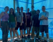 Eine Gruppe von sieben Personen steht in einem Raum der Hochschule Coburg mit blau getönten Fenstern. Sie sind leger gekleidet und lächeln in die Kamera. Das durchfallende Sonnenlicht durchflutet sie und verleiht dem glänzenden Boden eine lebendige Atmosphäre.