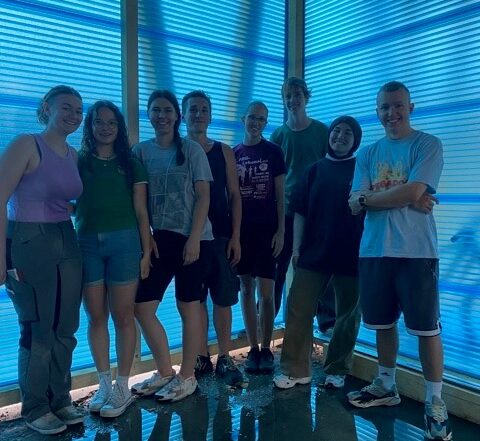 Eine Gruppe von sieben Personen steht in einem Raum der Hochschule Coburg mit blau getönten Fenstern. Sie sind leger gekleidet und lächeln in die Kamera. Das durchfallende Sonnenlicht durchflutet sie und verleiht dem glänzenden Boden eine lebendige Atmosphäre.