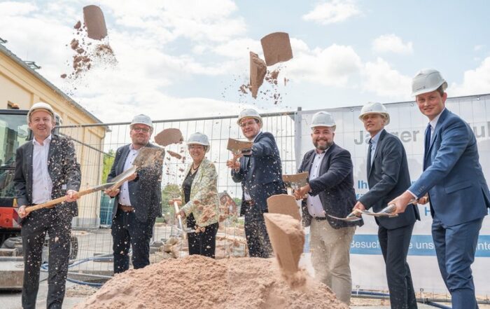 Eine Gruppe von sieben Personen in Business-Kleidung und mit Bauhelmen nimmt an einer Grundsteinlegungszeremonie an der Hochschule Coburg teil. Lächelnd schaufeln sie mit Schaufeln Erde in die Erde. Eine Baustelle und ein Bauzaun dienen als Kulisse.