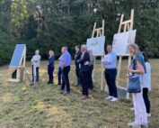 Eine Gruppe von Menschen steht auf einer Rasenfläche neben einer Ausstellung zu Solarmodulen und Informationstafeln an der Hochschule Coburg. Sie scheinen jemandem zuzuhören, der spricht. Im Hintergrund sind hohe Bäume zu sehen.