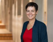 Eine Person mit kurzen dunklen Haaren, einem roten Oberteil und einer dunklen Jacke steht lächelt in einem sanft beleuchteten Flur der Hochschule Coburg. Im Hintergrund ist eine Treppe zu sehen.