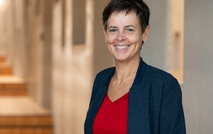 Eine Person mit kurzen dunklen Haaren, einem roten Oberteil und einer dunklen Jacke steht lächelt in einem sanft beleuchteten Flur der Hochschule Coburg. Im Hintergrund ist eine Treppe zu sehen.