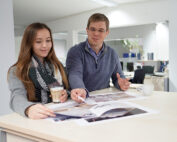 Ein Mann und eine Frau stehen an einem Tisch in einem Büro und besprechen die vor ihnen ausgebreiteten Dokumente. Die Frau, die eine Kaffeetasse der Hochschule Coburg in der Hand hält, unterhält sich mit dem Mann. Im Hintergrund sind sie von Bürogeräten und Pflanzen umgeben.
