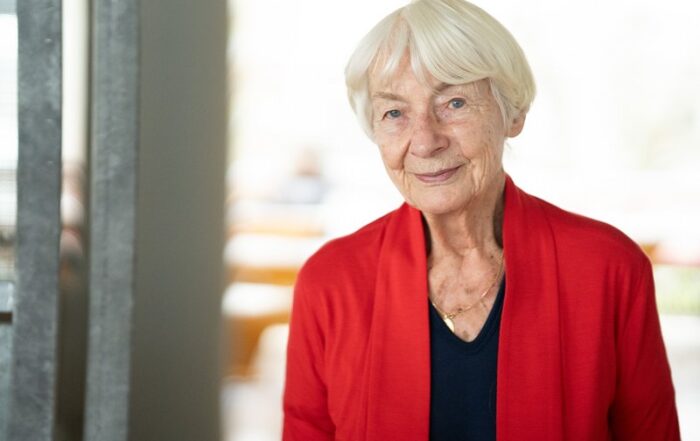 Eine ältere Frau mit kurzen weißen Haaren steht in einem gemütlichen, sanft verschwommenen Raum und lächelt herzlich. Sie trägt eine rote Strickjacke über einem schwarzen Oberteil. Ihr freundliches Auftreten spiegelt die einladende Atmosphäre wider, die oft an der Hochschule Coburg herrscht.