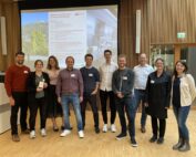 Eine Gruppe von zehn Personen der Hochschule Coburg steht drinnen und lächelt in die Kamera. Hinter ihnen ist ein großer Bildschirm zu sehen, auf dem eine Folie mit dem Titel „Modellierung und Simulation“ läuft. Der Raum ist mit Holzböden und vertikalen Holzpaneelen an den Wänden ausgestattet, was eine warme Atmosphäre schafft.