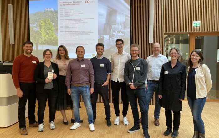 Eine Gruppe von zehn Personen der Hochschule Coburg steht drinnen und lächelt in die Kamera. Hinter ihnen ist ein großer Bildschirm zu sehen, auf dem eine Folie mit dem Titel „Modellierung und Simulation“ läuft. Der Raum ist mit Holzböden und vertikalen Holzpaneelen an den Wänden ausgestattet, was eine warme Atmosphäre schafft.