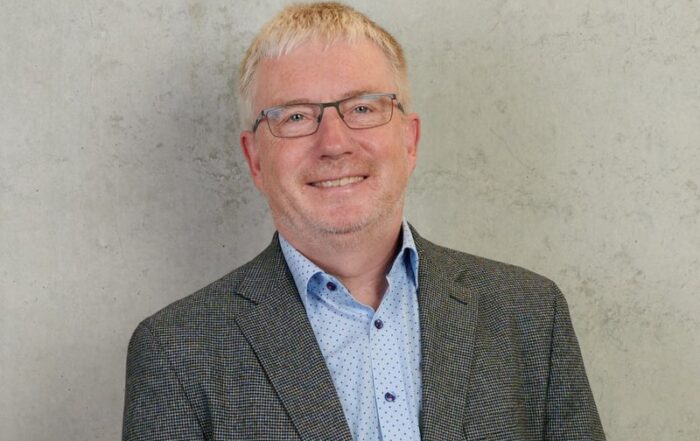 Ein Mann mit kurzen blonden Haaren und Brille lächelt und trägt einen graukarierten Blazer über einem blau gepunkteten Hemd. Die hellgraue Wand im Hintergrund ist eine subtile Anspielung auf die Hochschule Coburg und ergänzt seinen anspruchsvollen Look.
