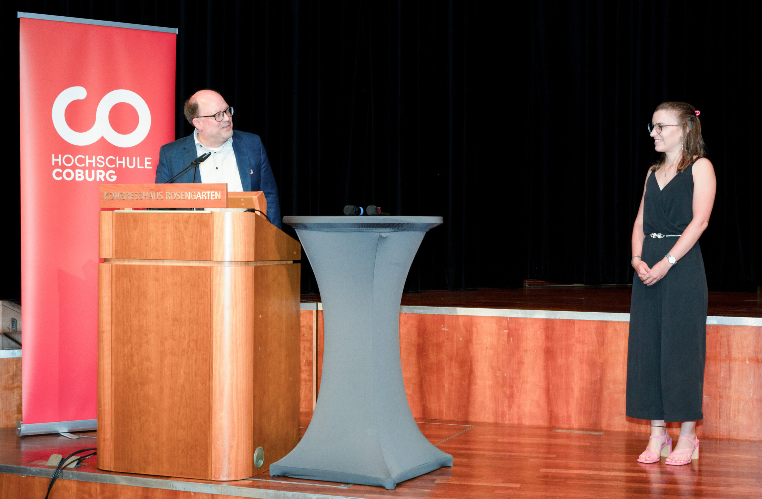 Ein Mann steht an einem Rednerpult mit einem Schild der Hochschule Coburg und spricht mit einer Frau in einem schwarzen Kleid auf der Bühne; zwischen ihnen steht ein hoher runder Tisch, im Hintergrund ist ein rotes Transparent zu sehen – während der Absolventenfeier 2024. at Hochschule Coburg