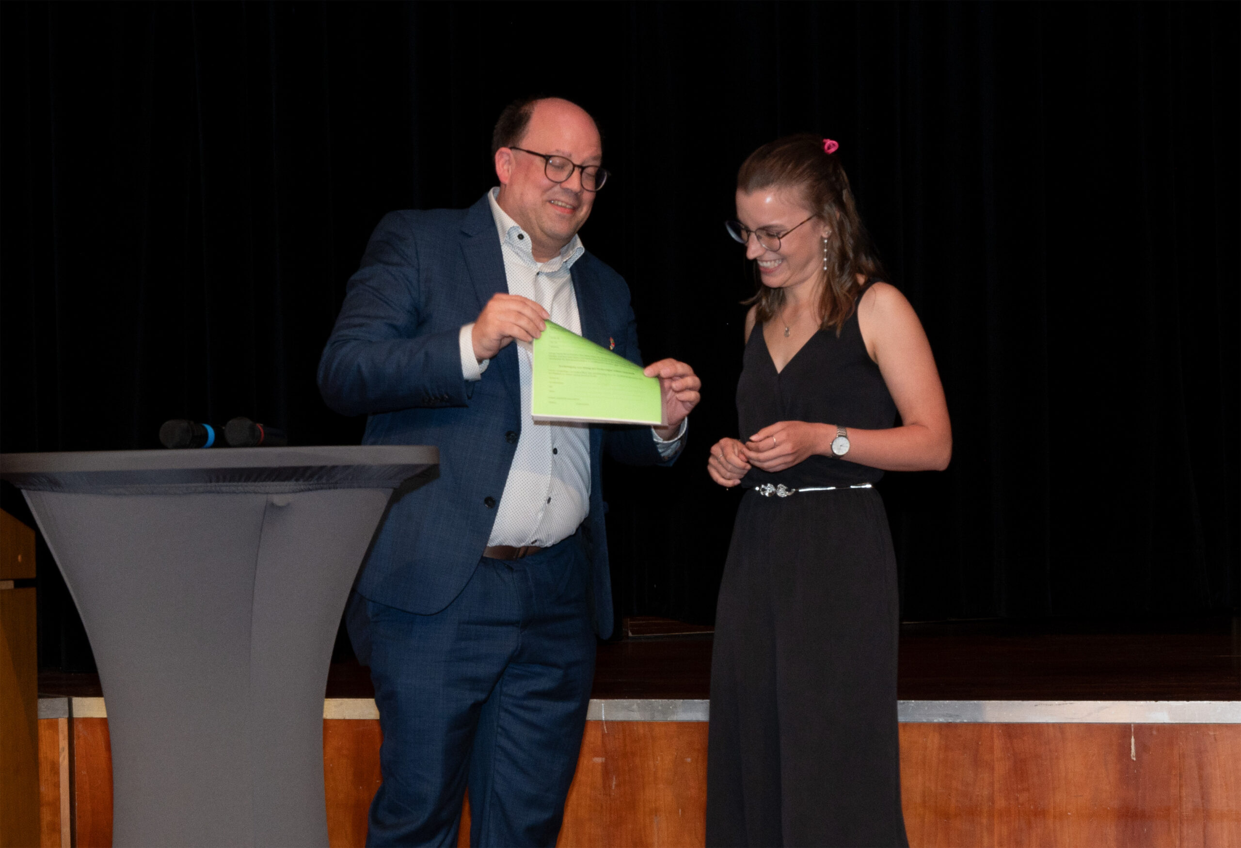 Bei der Absolventenfeier 2024 überreicht ein Mann im Anzug einer lächelnden Frau in einem schwarzen Kleid auf der Bühne neben einem kleinen runden Tisch vor einem dunklen Vorhang eine Urkunde – ein feierlicher Moment inmitten der lebhaften und bunten Feier. at Hochschule Coburg