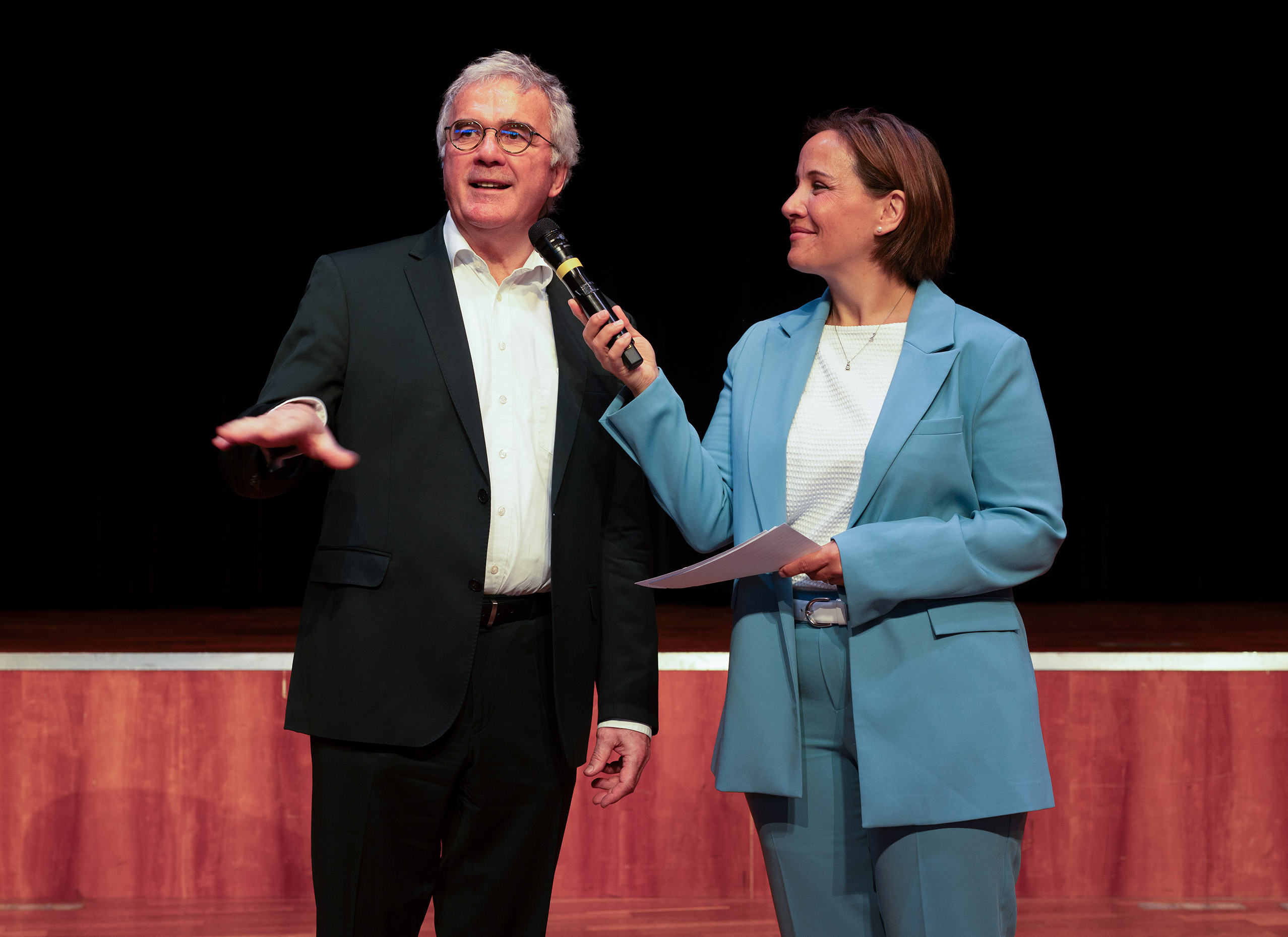 Bei der Absolventenfeier 2024 hält ein Mann in einem schwarzen Anzug eine Rede, während eine Frau in einem hellblauen Anzug das Mikrofon hält und ihn auf der Bühne vor einem dunklen Hintergrund anlächelt, wodurch eine feierliche und zugleich bunte Atmosphäre entsteht. at Hochschule Coburg