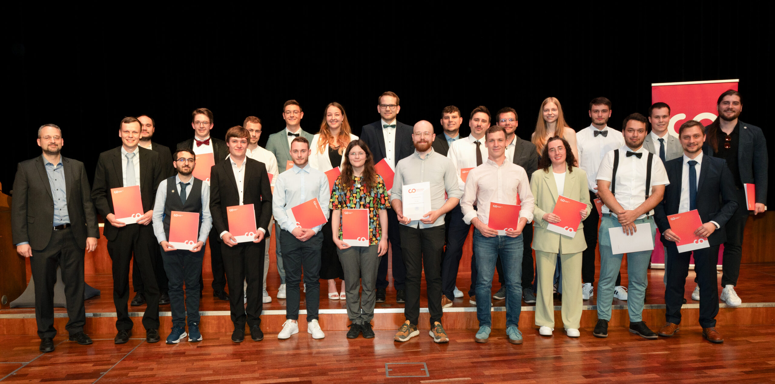 Eine Gruppe von 23 Personen, von denen einige ihre Zeugnisse in den Händen halten, posiert gemeinsam auf einer Holzbühne vor einem dunklen Hintergrund. Die meisten sind in Business- oder halbformeller Kleidung gekleidet, lächeln in die Kamera und fangen so die feierliche Atmosphäre der Absolventenfeier 2024 ein. at Hochschule Coburg