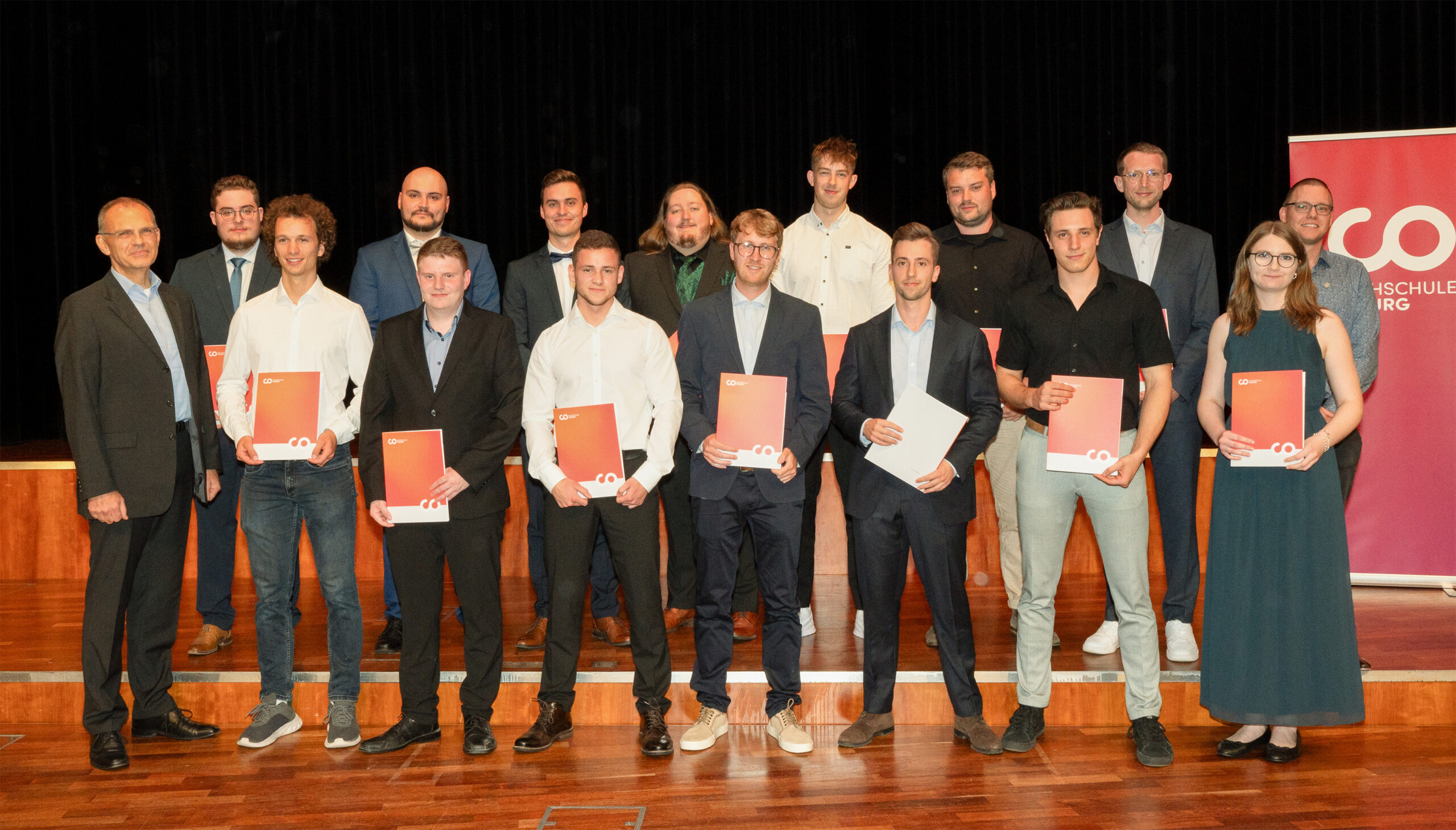 Eine Gruppe von Menschen in festlicher Kleidung steht auf einer Bühne, hält Urkunden in den Händen und lächelt für ein Foto während der feierlichen Abschlussfeier 2024. Im Hintergrund ist ein rotes Transparent mit weißer Schrift zu sehen. at Hochschule Coburg