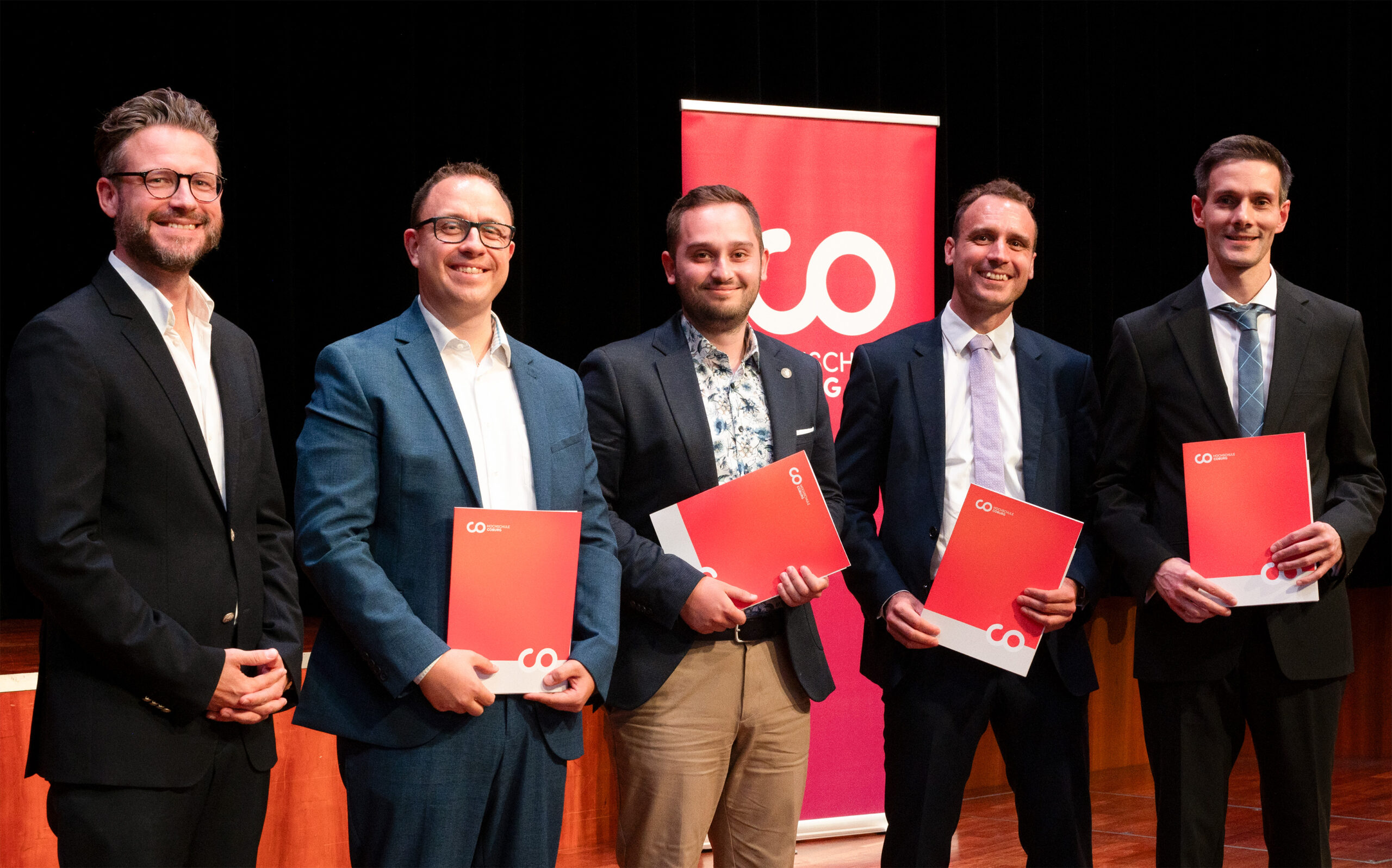 Fünf Männer in Anzügen stehen im Innenraum, lächeln und halten rote Mappen in den Händen, hinter ihnen auf einer Bühne hängt ein feierliches rot-weißes Banner – ein stolzer Moment der Absolventenfeier 2024. at Hochschule Coburg
