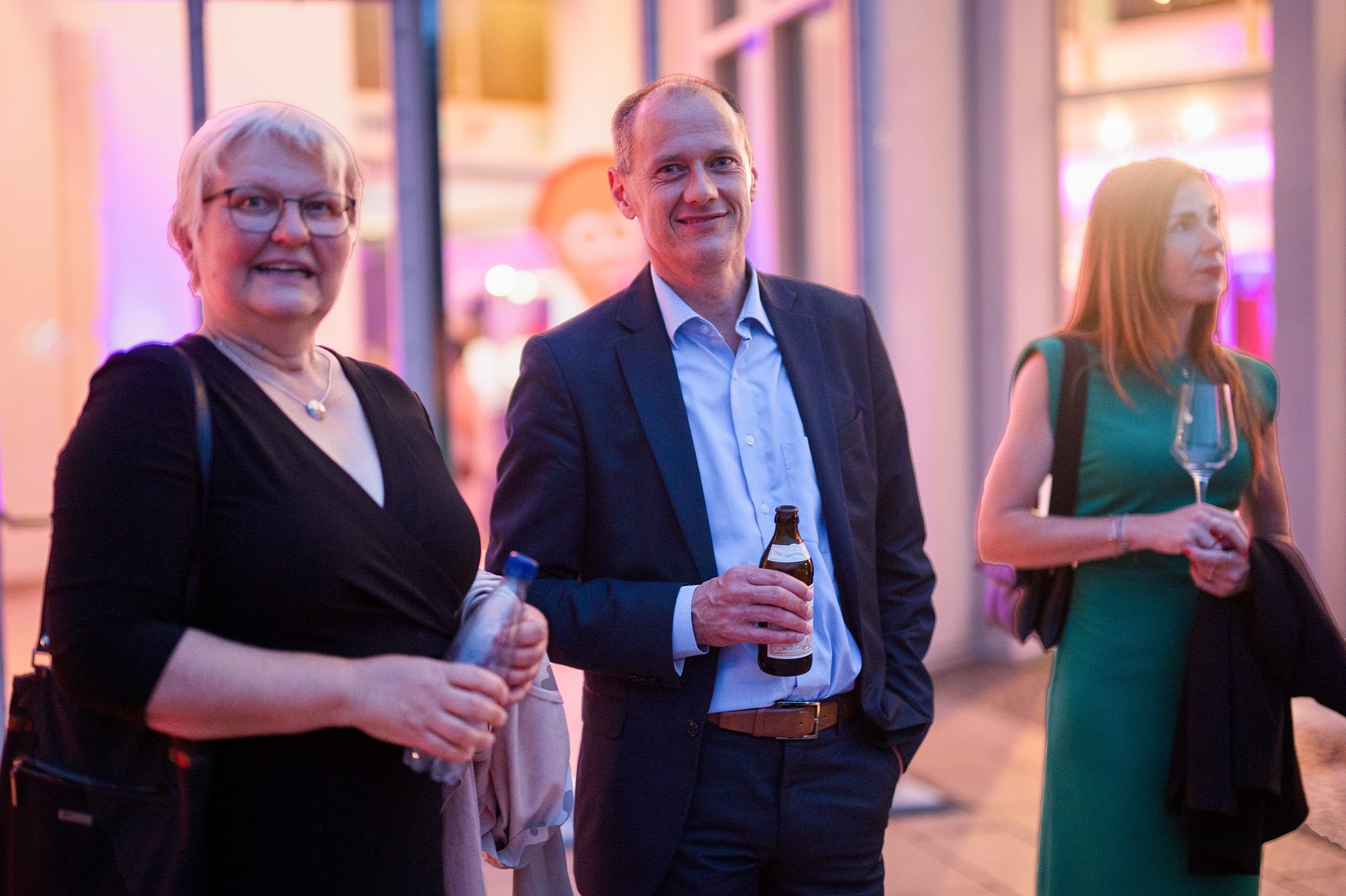 Drei elegant gekleidete Erwachsene stehen drinnen bei einer feierlichen Abschlussfeier 2024, halten Getränke in der Hand und lächeln. Der Hintergrund ist sanft in bunten Rosa- und Violetttönen beleuchtet. at Hochschule Coburg