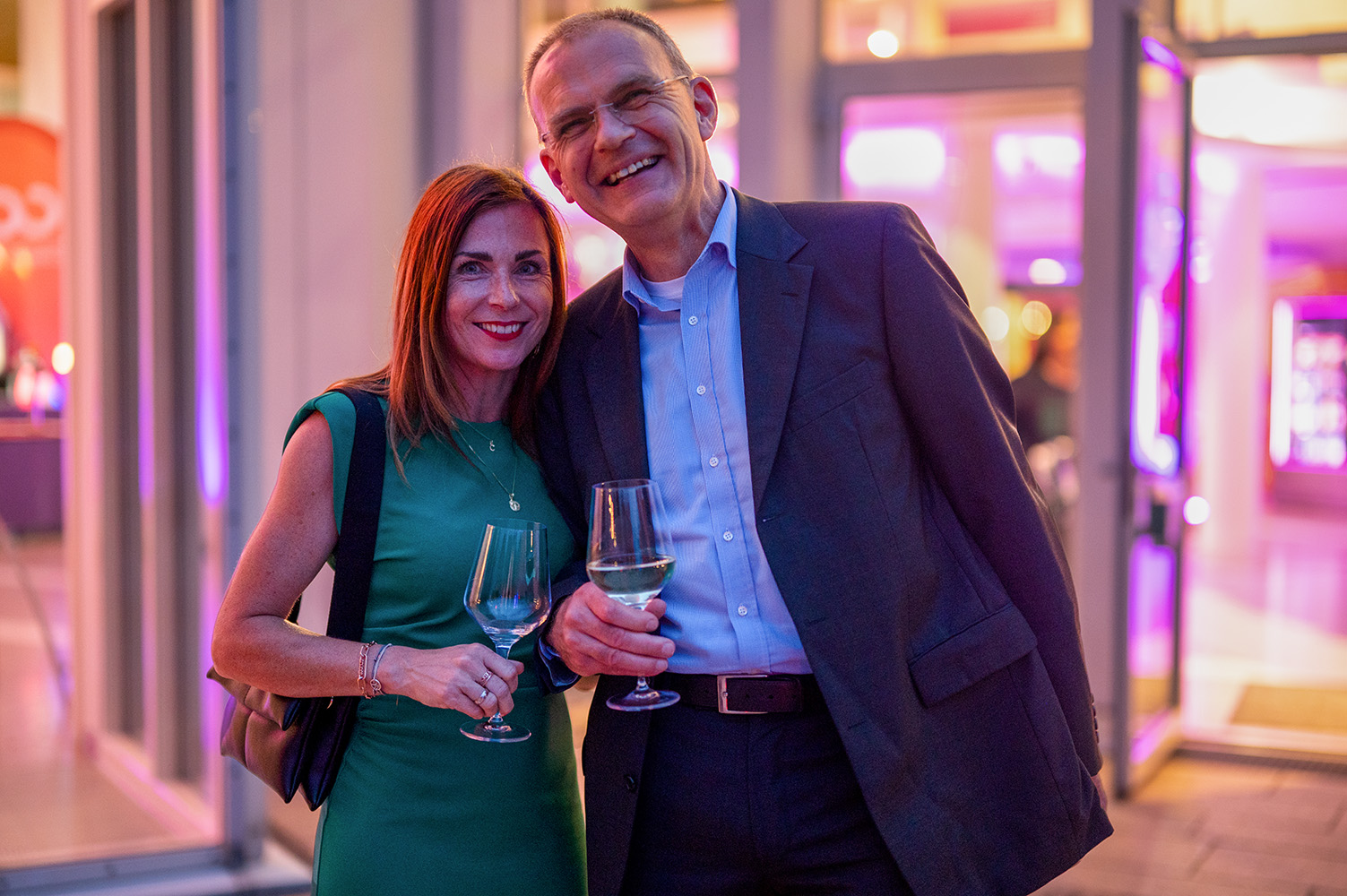 Eine Frau in einem grünen Kleid und ein Mann in einem dunklen Anzug lächeln, während sie bei einer Absolventenfeier 2024 in einem Saal, der von leuchtendem violettem Licht erhellt wird, Weingläser in den Händen halten. at Hochschule Coburg