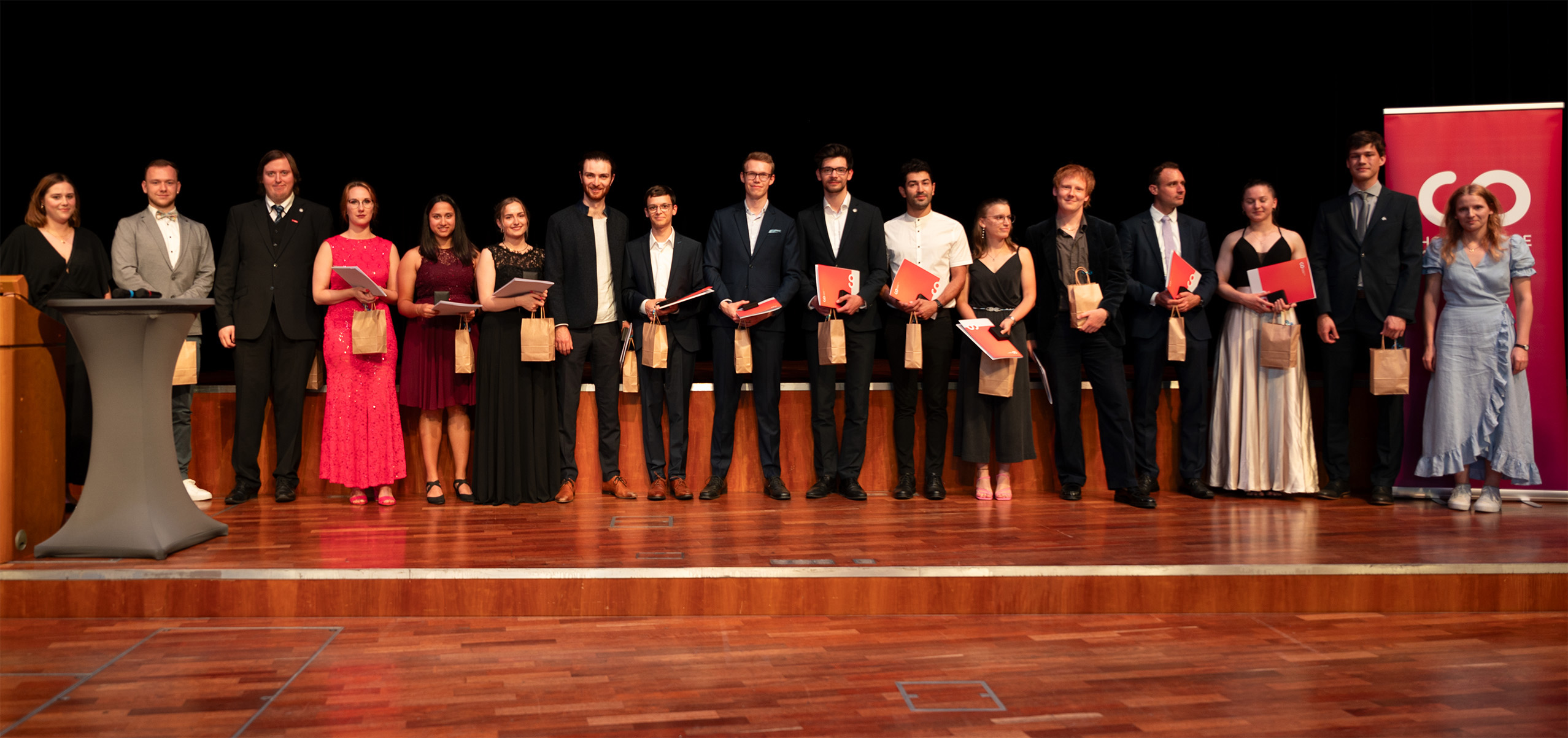 Eine Gruppe von 16 Personen in festlicher Kleidung steht auf einer Bühne, hält Geschenktüten und Urkunden in den Händen und lächelt für ein Foto während der feierlichen Abschlussfeier 2024. Auf der rechten Seite des Bildes ist ein rot-weißes Banner zu sehen. at Hochschule Coburg