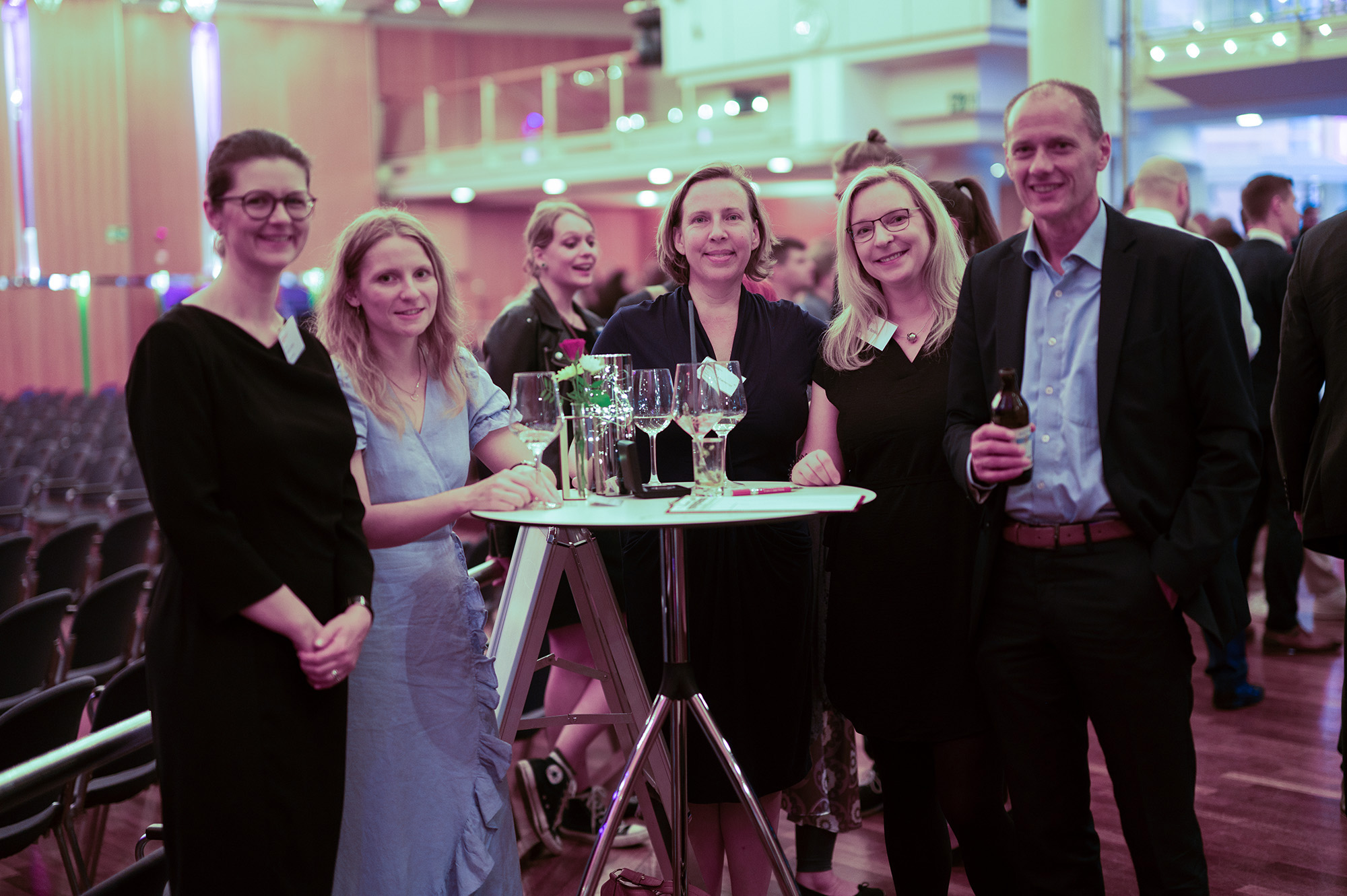 Bei einer feierlichen Abschlussfeier im Jahr 2024 stehen fünf Personen bei einer formellen Veranstaltung in einem großen, hell erleuchteten Saal um einen hohen Tisch mit leeren Weingläsern und einer Flasche herum. Im Hintergrund sind weitere Gäste zu sehen. at Hochschule Coburg