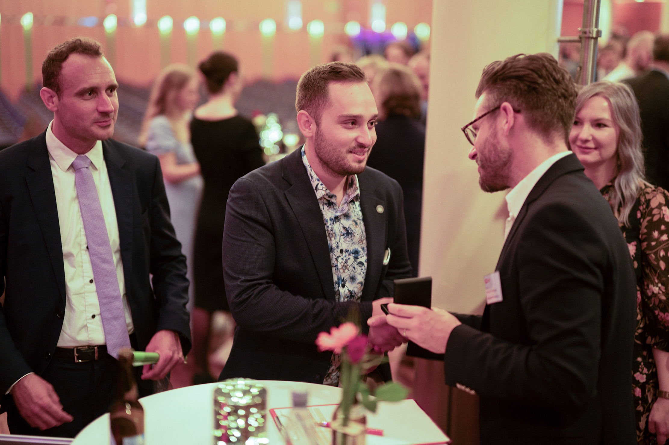 Vier Personen in festlicher Kleidung unterhalten sich an einem runden Tisch, auf dem Getränke und Blumen stehen, bei einer Absolventenfeier 2024 in einem Saal; zwei Männer in der Mitte geben sich die Hand, während die anderen zuschauen und lächeln. at Hochschule Coburg