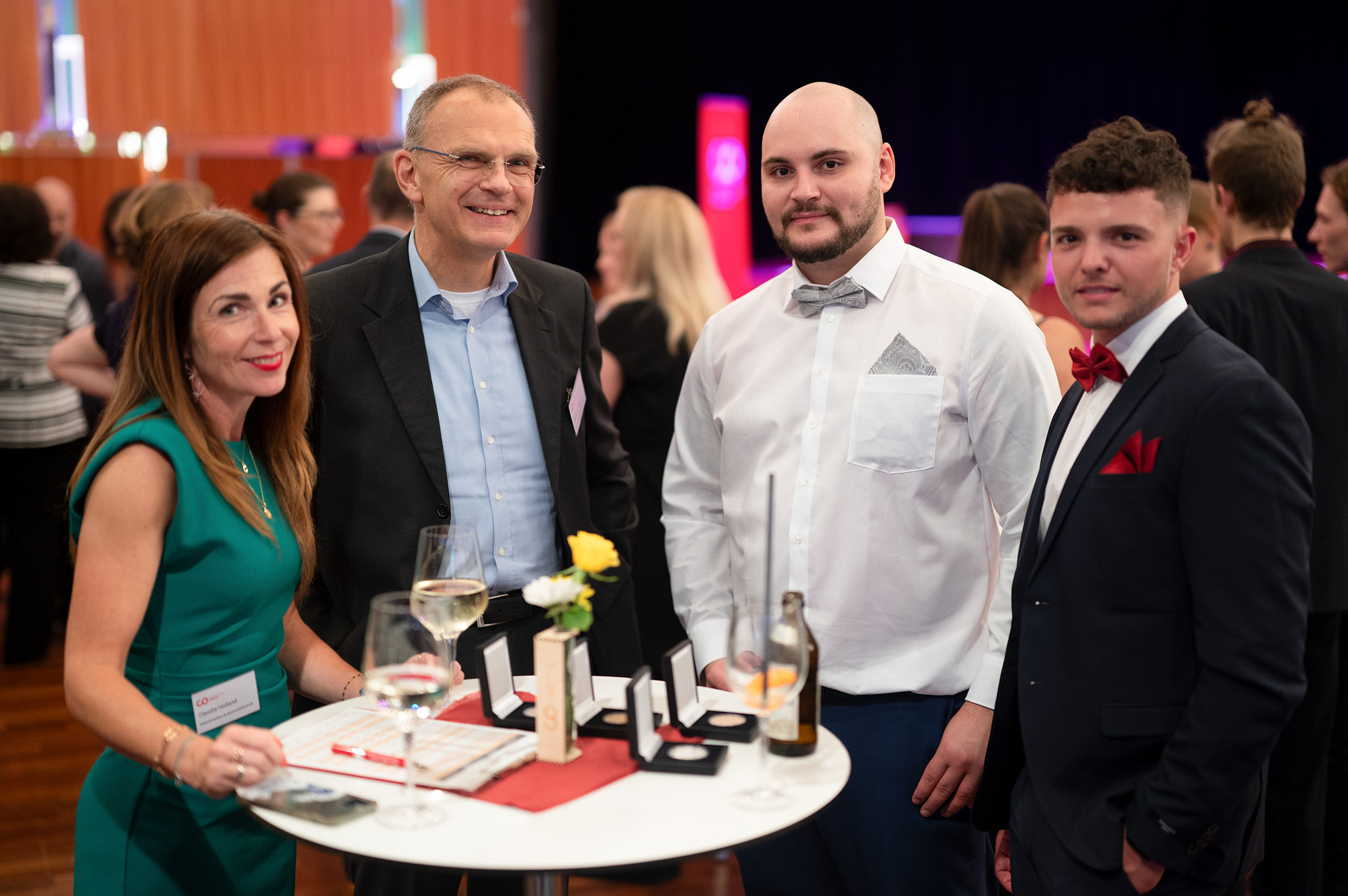 Vier Personen in festlicher Kleidung stehen bei einer feierlichen Abschlussfeier 2024 um einen kleinen Tisch herum, auf dem Getränke, Blumen und Schachteln stehen. Im Hintergrund sind weitere Gäste und die Dekoration der Veranstaltung zu sehen. at Hochschule Coburg