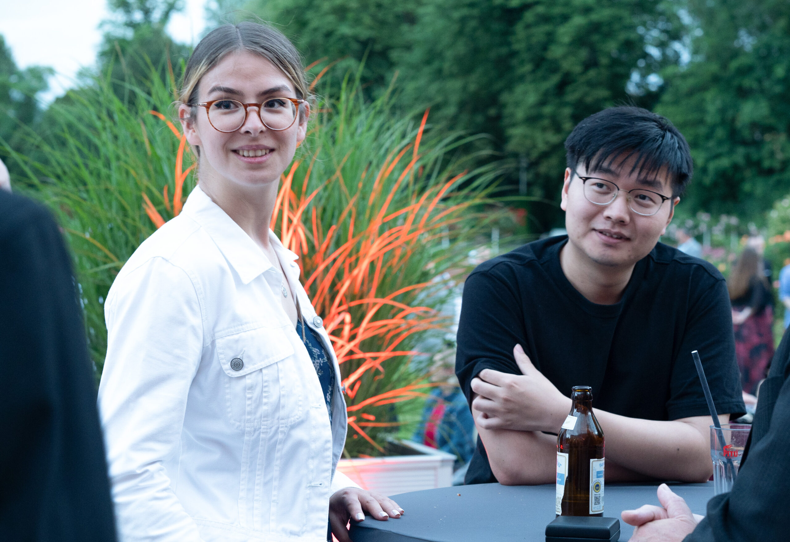 Zwei Personen sitzen während einer feierlichen Abschlussfeier 2024 an einem Tisch im Freien. Die Frau mit Brille und weißer Jacke lächelt in die Kamera, während der Mann mit Brille und schwarzem Hemd mit einem Getränk neben ihr sitzt. Im Hintergrund sind grüne Pflanzen und orangefarbene Lichter zu sehen. at Hochschule Coburg