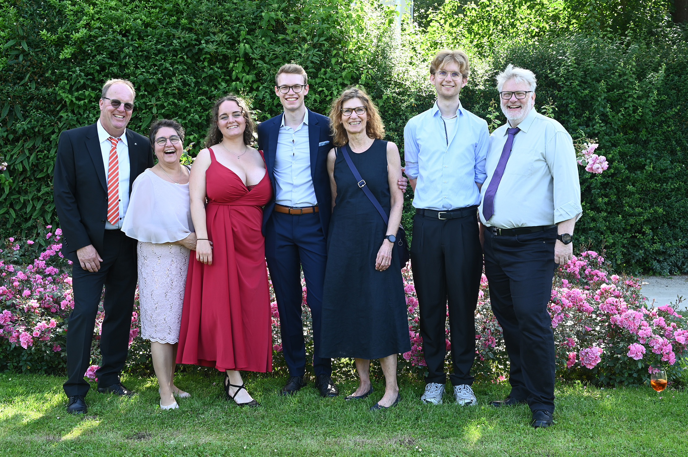Sieben Erwachsene stehen lächelnd in einer Reihe vor grünen Büschen und rosa Blumen, gekleidet in festliche, formelle und halbformelle Kleidung, und genießen die sonnige Absolventenfeier 2024 im Freien. at Hochschule Coburg