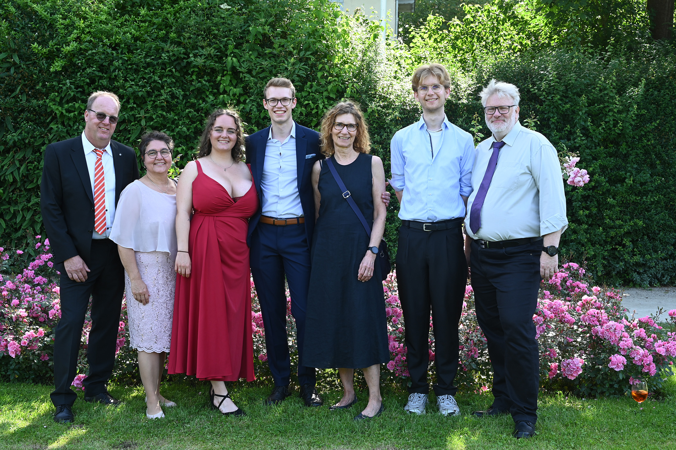 Sieben Personen posieren und lächeln an einem sonnigen Tag in formeller und halbformeller Kleidung vor blühenden rosa Blumen und grünen Hecken und halten so einen feierlichen Moment bei der Absolventenfeier 2024 fest. at Hochschule Coburg