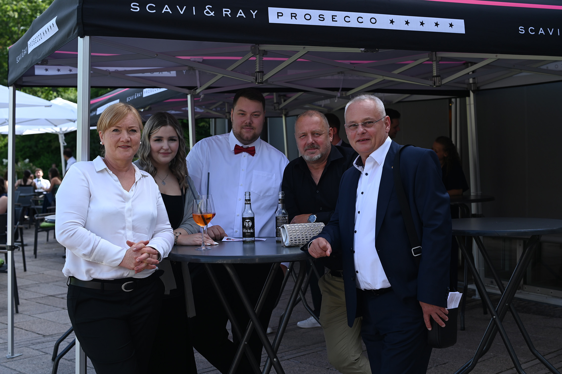 Fünf Erwachsene stehen im Freien unter einem Zeltdach bei der feierlichen Abschlussfeier 2024 um einen hohen Tisch herum; einige halten Getränke in der Hand, alle sind elegant oder im Business-Casual-Stil gekleidet und posieren lächelnd für die Kamera. at Hochschule Coburg