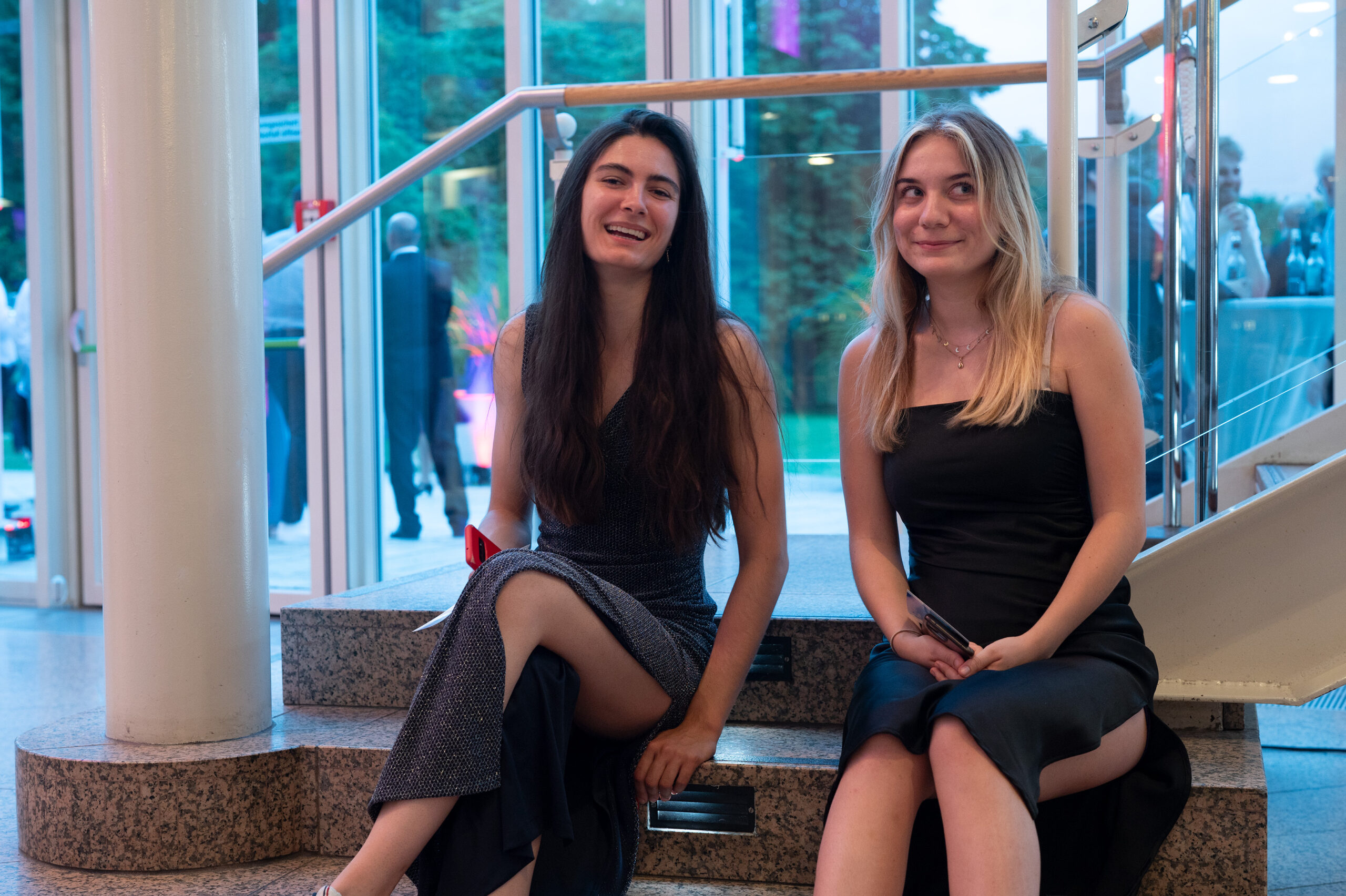 Zwei junge Frauen in Abendkleidern sitzen auf einer Innentreppe und lächeln während einer feierlichen Abschlussfeier. Durch große Glastüren und Fenster sind im Hintergrund ein Garten und feiernde Menschen zu sehen. Die Atmosphäre ist hell und festlich. at Hochschule Coburg