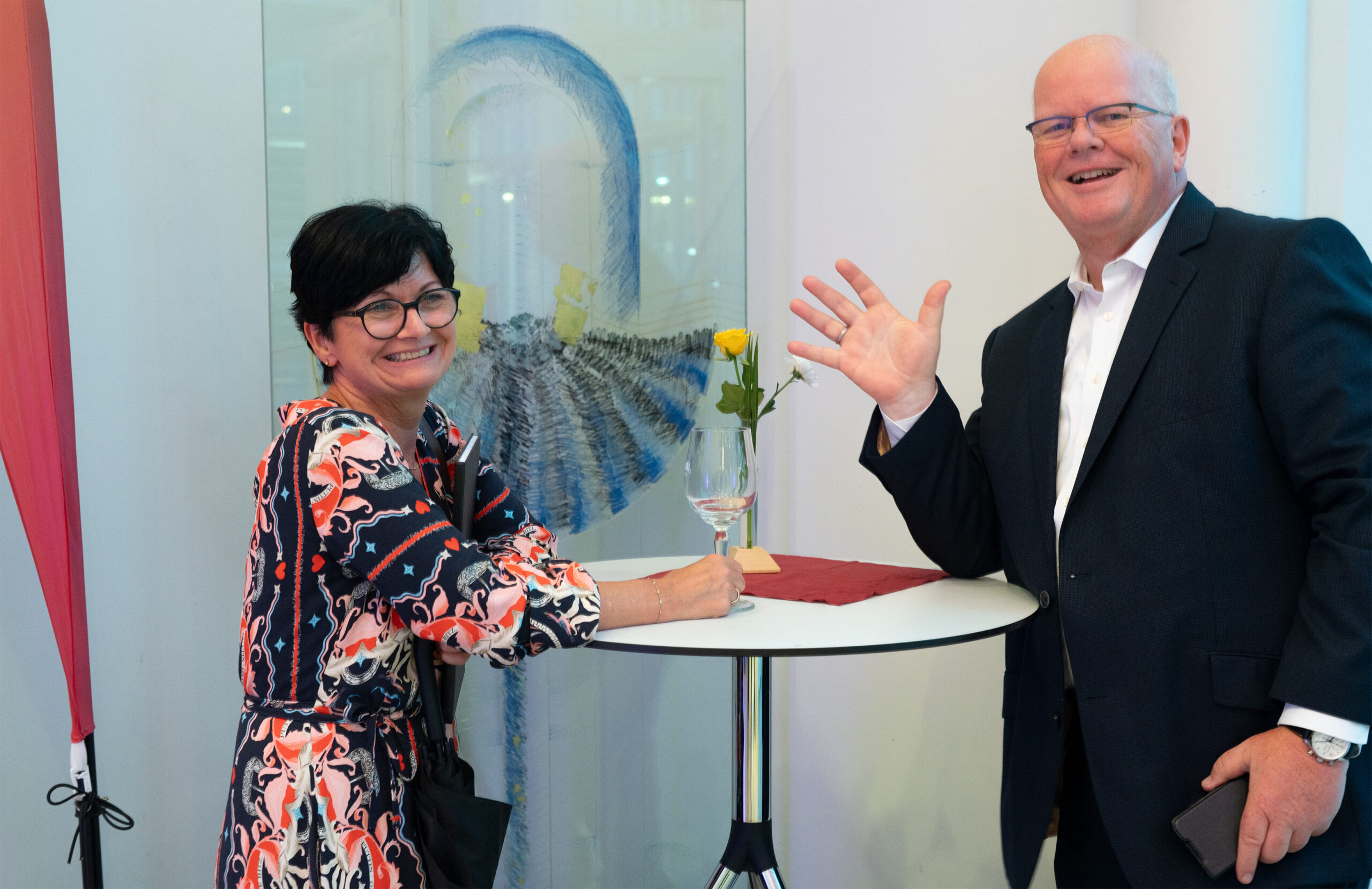 Eine Frau und ein Mann stehen lächelnd an einem hohen runden Tisch, auf dem eine gelbe Rose in einer Vase und ein leeres Weinglas stehen, und genießen gemeinsam einen feierlichen Moment. Der Mann winkt in die Kamera und fängt so die fröhliche Stimmung der Absolventenfeier 2024 ein. Im Hintergrund ist ein abstraktes Gemälde zu sehen. at Hochschule Coburg
