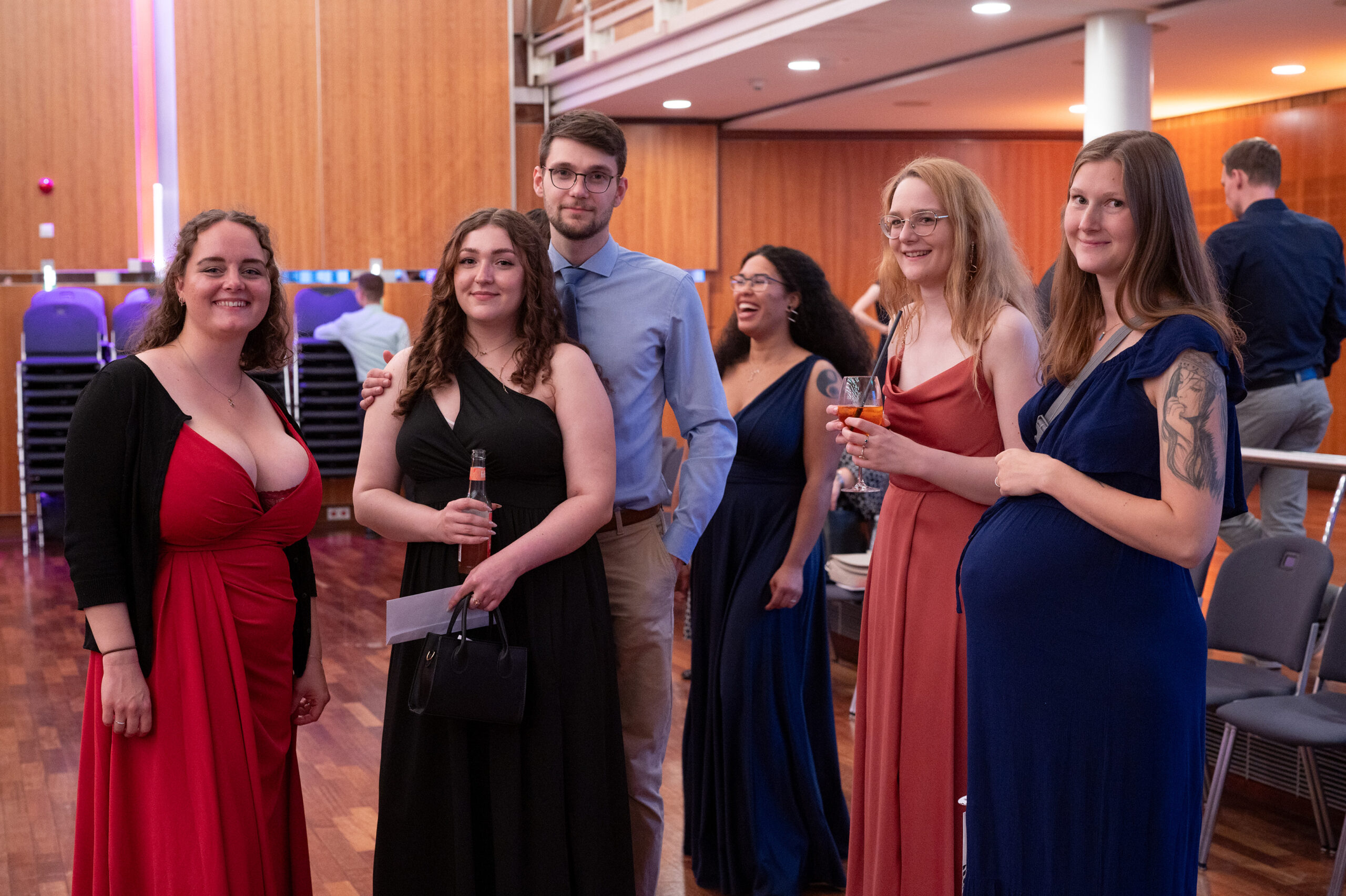 Sechs elegant gekleidete Personen stehen bei einer Veranstaltung in einem Saal beieinander und lächeln in die Kamera. Die Frauen tragen farbenfrohe Kleider, und eine von ihnen hält ein Getränk in der Hand. Der Schauplatz scheint ein Saal mit Holzboden und gestapelten Stühlen zu sein, was eine feierliche Atmosphäre schafft, wie sie zu einer Absolventenfeier 2024 passt. at Hochschule Coburg