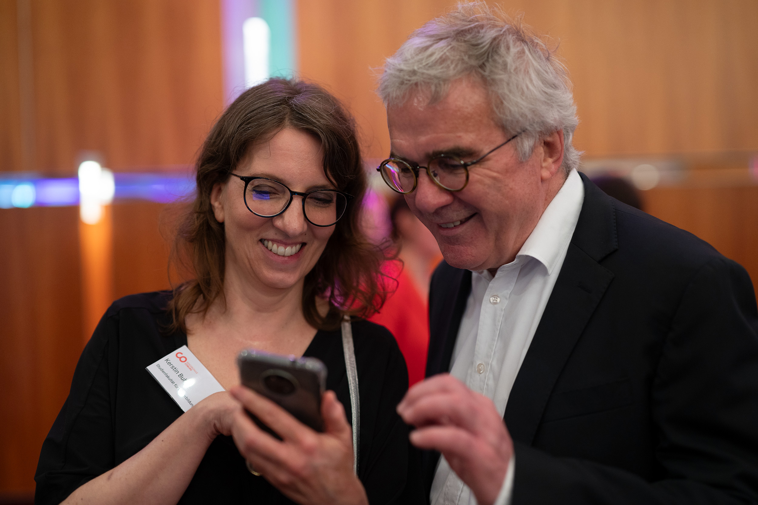 Zwei Menschen mit Brille und in Business-Kleidung lächeln, während sie gemeinsam in einem hell beleuchteten Raum mit Holzwänden auf ein Smartphone blicken – vielleicht teilen sie einen ungezwungenen Moment von der kürzlich stattgefundenen Absolventenfeier 2024. at Hochschule Coburg