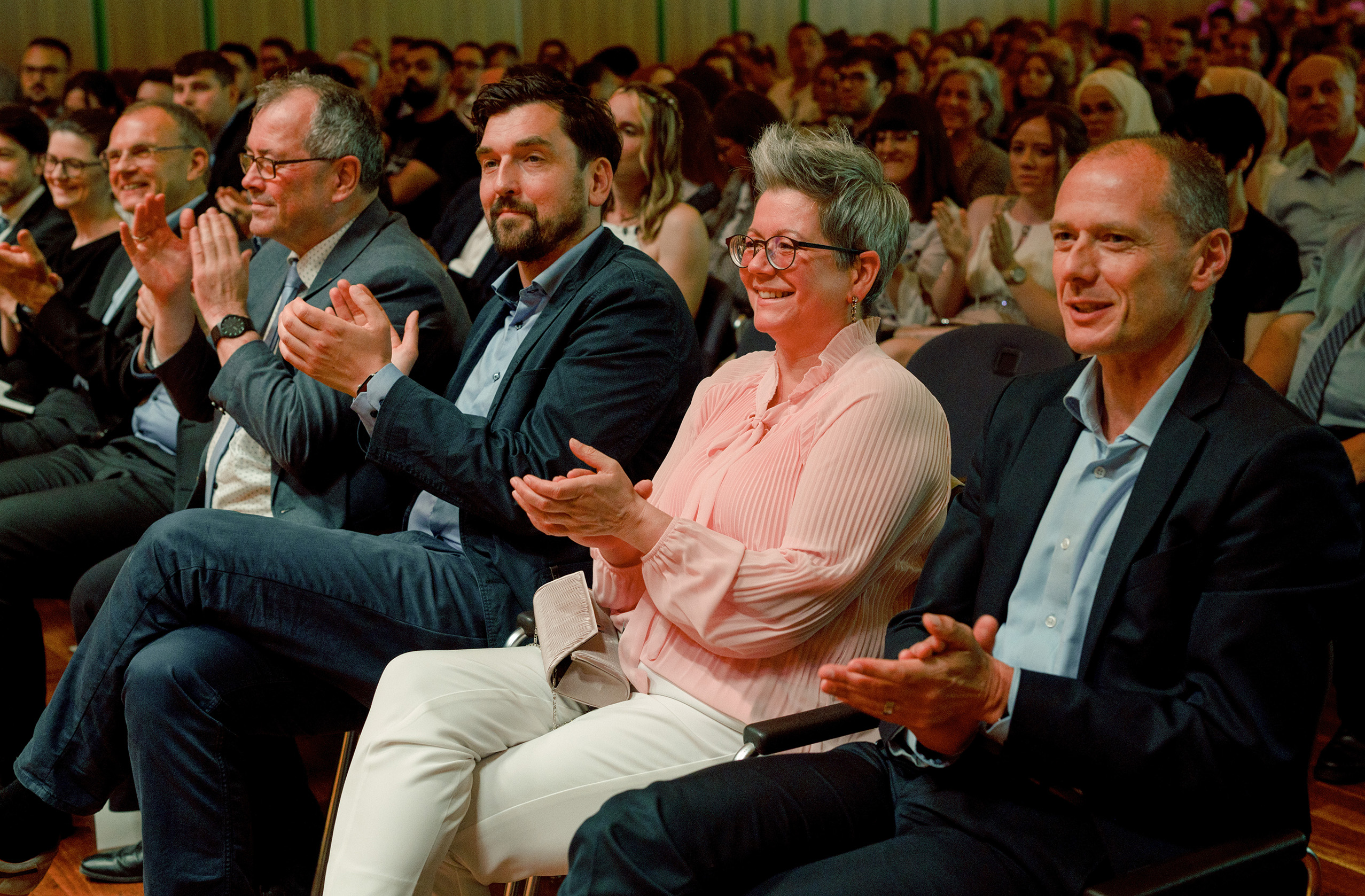 Ein großes, feierliches Publikum sitzt bei einer Veranstaltung in einem Saal und klatscht. Im Vordergrund sitzen vier Personen in Geschäftskleidung in einer Reihe, lächeln und applaudieren bei der bunten Absolventenfeier 2024; hinter ihnen sind weitere Gäste zu sehen. at Hochschule Coburg