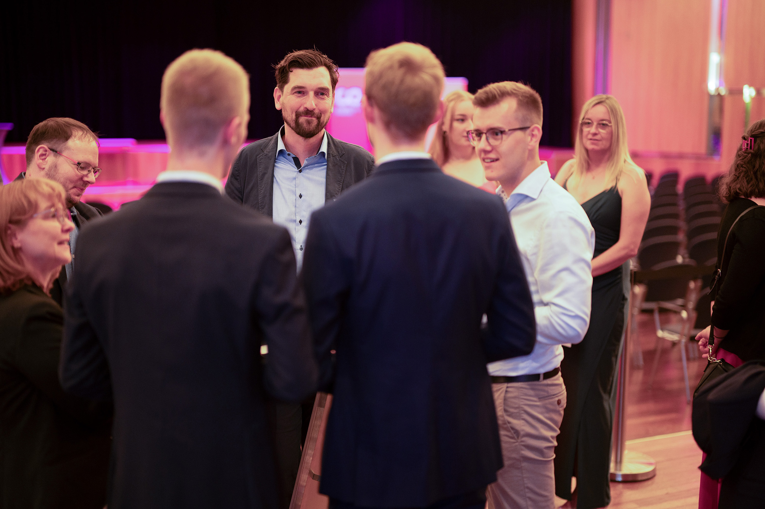 Eine Gruppe von Menschen in formeller und halbformeller Kleidung steht im Kreis und unterhält sich in einem großen, warm beleuchteten Veranstaltungsraum, hinter dem Reihen leerer Stühle zu sehen sind, was eine feierliche Atmosphäre schafft, wie sie zu einer Absolventenfeier 2024 passt. at Hochschule Coburg