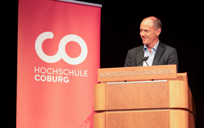 Ein Mann im Anzug steht an einem hölzernen Rednerpult mit der Aufschrift „KONGRESSHAUS ROSENGARTEN“ und hält eine feierliche Rede, im Hintergrund ist ein rotes Banner mit der Aufschrift „HOCHSCHULE COBURG“ zu sehen – ein Bild, das den Geist der Absolventenfeier 2024 einfängt. at Hochschule Coburg