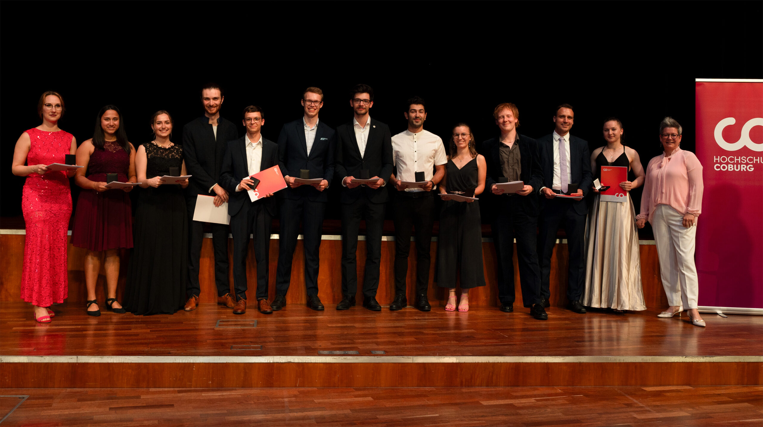 Dreizehn Personen in festlicher Kleidung stehen in einer Reihe auf der Bühne, einige halten Urkunden in der Hand, vor einem Banner der Hochschule Coburg auf der rechten Seite und lächeln für ein Gruppenfoto bei der feierlichen Absolventenfeier 2024. at Hochschule Coburg
