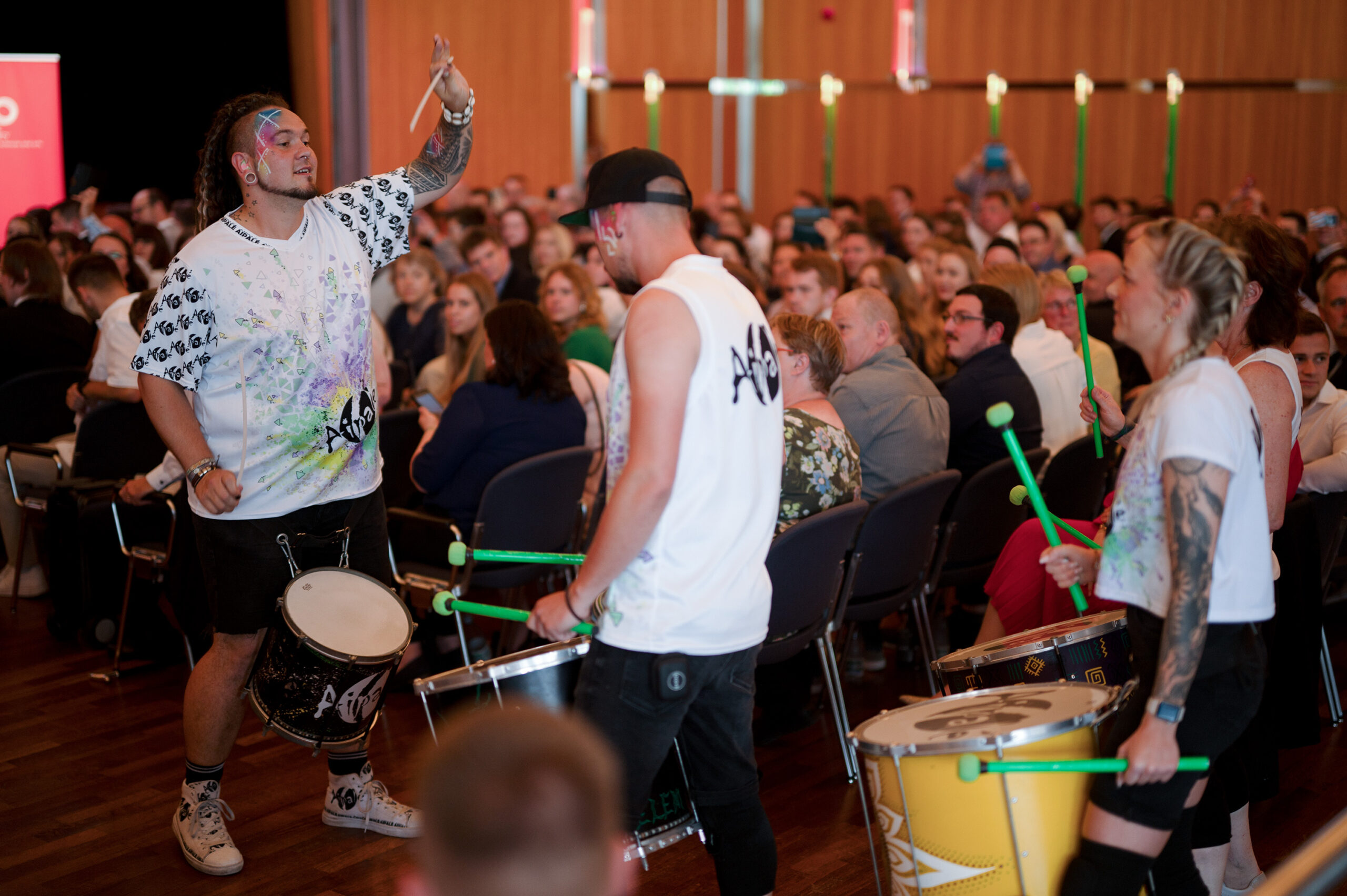 Drei Schlagzeuger treten während der Absolventenfeier 2024 in einem holzgetäfelten Raum vor einem sitzenden Publikum energiegeladen auf. Die Musiker tragen einheitliche Hemden und spielen auf bunten Trommeln; mit ihrer lebhaften Darbietung begeistern sie das Publikum und verleihen der Feier einen unvergesslichen Rhythmus. at Hochschule Coburg