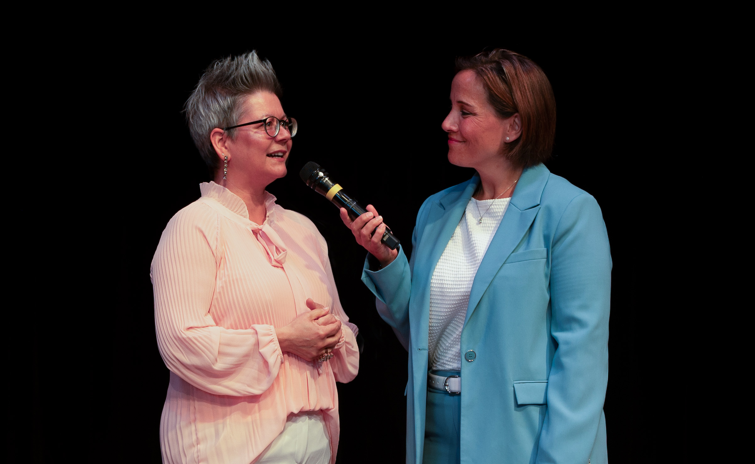 Zwei Frauen stehen während der Absolventenfeier 2024 auf der Bühne. Die eine trägt eine hellrosa Bluse und eine Brille und spricht, während die andere, die einen blauen Anzug trägt, ihr mit strahlendem Lächeln ein Mikrofon entgegenhält. Der Hintergrund ist schwarz, wodurch die Szene noch bunter und lebendiger wirkt. at Hochschule Coburg