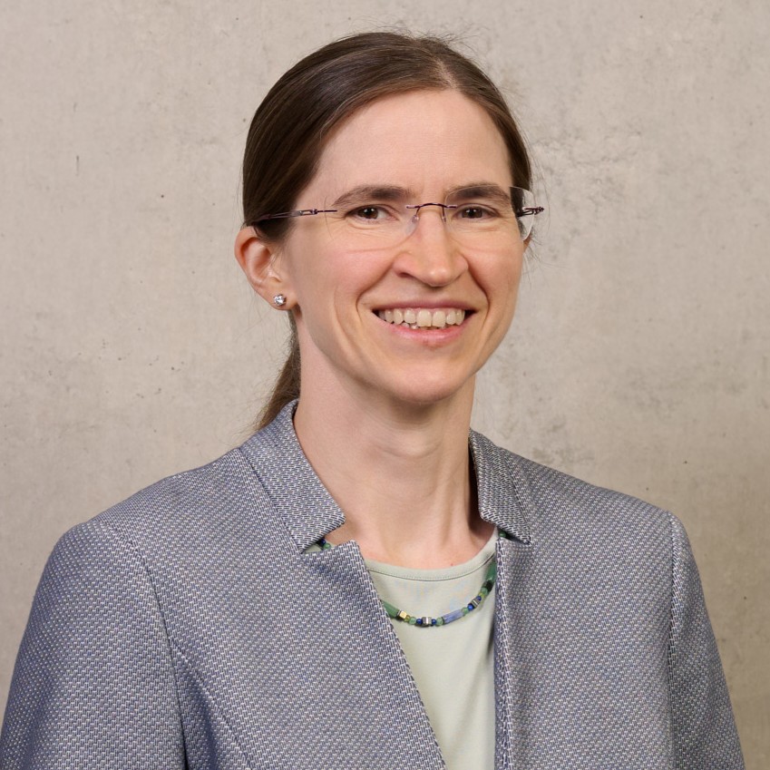 Eine Frau mit Brille und langen, nach hinten gebundenen braunen Haaren, die einen grauen Blazer, ein helles Oberteil und eine Perlenkette trägt, lächelt vor einem schlichten, hellen Hintergrund – das ist Bettina Friedel. at Hochschule Coburg