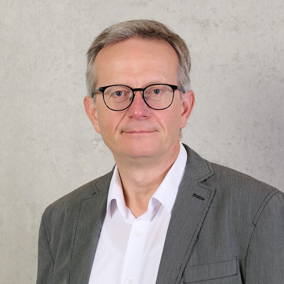 Prof. Dr. Olaf Huth, a middle-aged man with short gray hair and glasses, wears a white shirt and gray suit jacket as he stands in front of a light gray textured background. at Hochschule Coburg