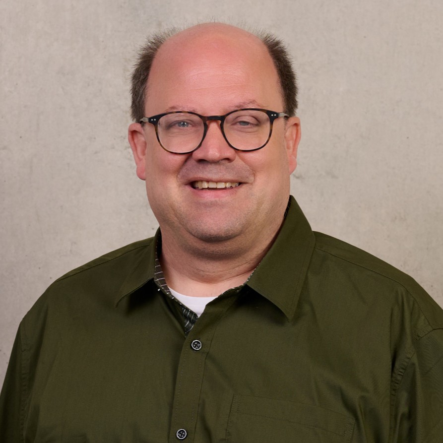 Prof. Dr. Mirko Kraft, ein lächelnder Mann mit Brille, kurzen braunen Haaren und einem olivgrünen Hemd, steht vor einem schlichten, hellen Hintergrund. at Hochschule Coburg