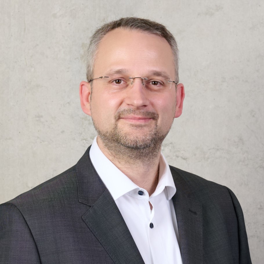 Prof. Dr. Jochen Merhof, a man with short gray hair, glasses, and a trimmed beard, is wearing a dark suit jacket over a white dress shirt and stands in front of a plain light-colored wall, smiling slightly at the camera. at Hochschule Coburg