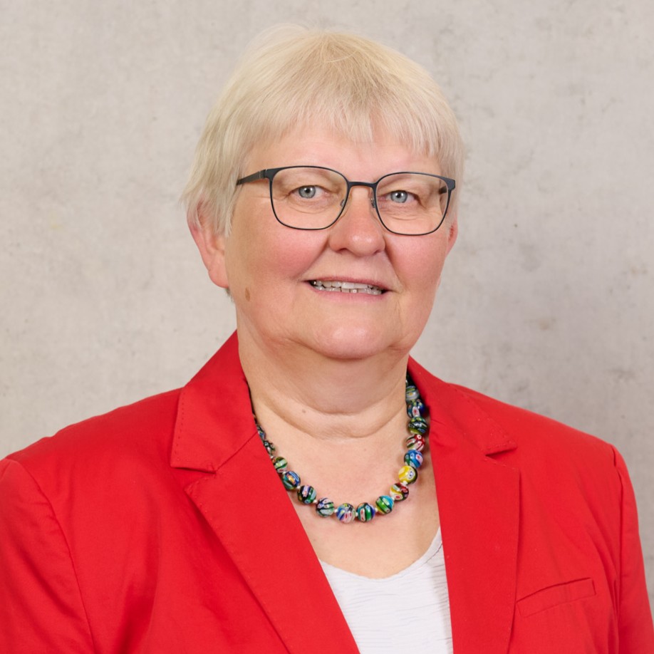 Prof. Dr. Jutta Michel, eine ältere Frau mit kurzen weißen Haaren, Brille und einer bunten Perlenkette, die einen roten Blazer und ein weißes Oberteil trägt, lächelt vor einem hellgrauen Hintergrund. at Hochschule Coburg
