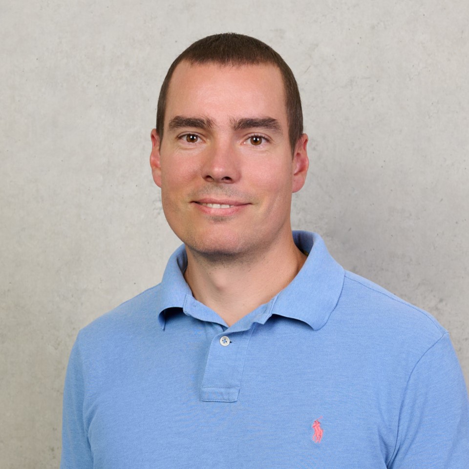 Roman Rischke, a man with short brown hair, wearing a light blue polo shirt, stands in front of a light gray textured wall, smiling slightly at the camera. at Hochschule Coburg