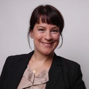 A woman with dark hair in a bun, wearing a black blazer and beige blouse, smiles at the camera against a plain light background. She has a pendant necklace and small stud earrings. at Hochschule Coburg