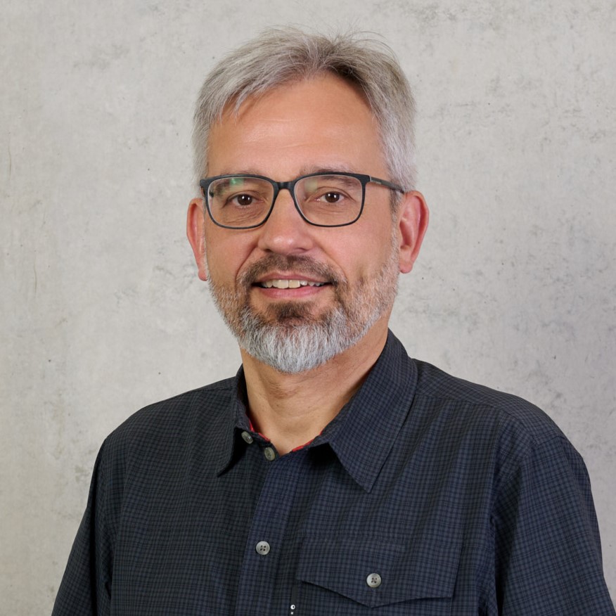 A middle-aged man with gray hair, glasses, and a beard is smiling. He is wearing a dark, checkered button-up shirt and standing in front of a light gray, textured background. at Hochschule Coburg