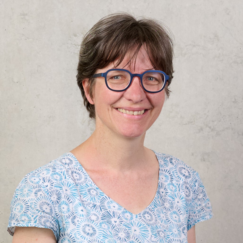 Alexandra Troi, eine Frau mit kurzen braunen Haaren und blauer Brille, lächelt in die Kamera. Sie trägt eine hellblau gemusterte Bluse und steht vor einer schlichten, hellen Wand. at Hochschule Coburg